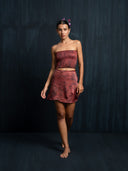 A woman stands barefoot against a dark background, wearing a strapless maroon crop top and matching wrap skirt, with her hair tied up and decorated with pink flowers.