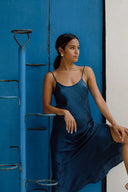 A woman in a silky blue slip dress stands beside a blue metal staircase and blue door, leaning casually with one hand on her knee and looking to the side.