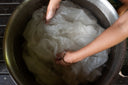 a close-up of a person’s hands submerging a piece of white fabric in a large metal basin filled with water. The person appears to be washing or soaking the fabric, possibly as part of a dyeing or cleaning process.
