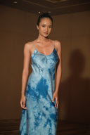 A person stands confidently in the Lily Silk Dress - Cosmic Indigo Tiedye from Rimmba | Sustainable Clothing, showcasing delicate spaghetti straps. The softly lit, neutral background enhances the elegance of this naturally dyed silk dress.