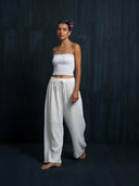 Against a dark background, a woman with flowers in her hair wears Rimmba The Brand’s Akasha Cotton Pants in white, paired with a strapless smocked top. She stands barefoot, calm, with one hand by her side and the other relaxed.