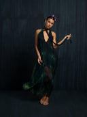 A barefoot woman poses against a dark backdrop in the Rimmba Aphrodite Dress - Aurora, a flowing sheer black and green open-back dress with a deep V-neck. She wears pink hair clips and holds a long black ribbon.