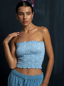 Woman wearing a baby indigo cotton embroidery strapless top and long pants against a dark background.
