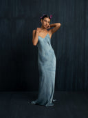A woman in a flowing Lily Silk Dress - Cosmic Indigo Tiedye by Rimmba | Sustainable Clothing stands against a dark background, eyes closed, with one arm behind her head and small pink flowers in her hair.