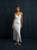 A woman stands barefoot against a dark backdrop, wearing the Lily White Silk Dress by Rimmba The Brand—long, with thin straps. A flower adorns her hair, and she has a calm expression.