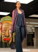 A model walks the runway in loose-fitting Nindy Silk Pant - Indigo by Rimmba | Sustainable Clothing, paired with a sheer purple scarf. Behind her is a colorful painting of a face with exaggerated features and a checkered headpiece.