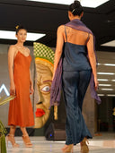 Two models walk the runway during a fashion show. One wears an orange slip dress, while the other showcases Rimmba’s Nindy Silk Pant in Indigo—crafted from mulberry silk and botanical dyes—with a purple scarf. A decorative mask is seen in the background.