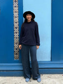 A person smiles at the camera in front of a blue wall with a decorative tile strip, wearing a black wide-brimmed hat, dark sweater, grey shoes, and Rimmba | Sustainable Clothing's Nindy Silk Pant - Indigo.