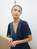 A woman in an indigo Olivia Vintage Silk Dress by Rimmba | Sustainable Clothing stands against a white textured wall, gazing thoughtfully to the side with her hands clasped and her hair in a bun with a gold accessory.