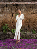 A woman in the Olivia Vintage Silk Dress - White by Rimmba | Sustainable Clothing stands barefoot on purple petals before a textured earthen wall, green plants growing at its base.