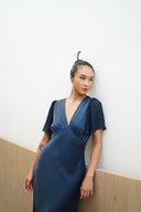 A person in a Rimmba Olivia Vintage Silk Dress - Indigo with flutter sleeves stands by a light textured wall. Their hair is styled in a bun, adorned with an accessory, showcasing tattoos on their arms.