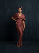 A woman stands barefoot against a dark background, wearing the Ria Silk Dress - Burgundy by Rimmba | Sustainable Clothing. Her hair is styled back with a flower behind her ear, and one hand rests on her hip.