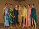 Seven women stand side by side in a row, wearing colorful and elegant dresses in various styles, posing confidently in a room with wooden floors and beige walls.