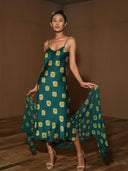 A woman stands indoors in the Rimmba Scarlette Silk Dress - Emerald Shibori, posing confidently on a wooden floor. The elegant mulberry silk dress is from Rimmba | Sustainable Clothing and features an emerald shibori print.
