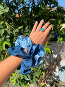 A hand showcases Rimmba | Sustainable Clothing Silk Scrunchies in navy and blue plus a blue gemstone ring, held up to green foliage and sunlight—highlighting gentle hair care with luxurious mulberry silk.