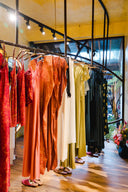 Colorful dresses and robes hang neatly on a rack in a boutique with wooden floors, large windows, and warm lighting. A bouquet of flowers sits on the floor to the right.