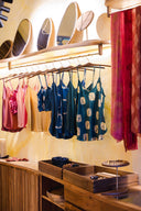 A clothing boutique display shows colorful sleeveless tops hanging on a rack beneath round mirrors. Jewelry and accessories are arranged on wooden trays below, with scarves hanging to the right.