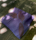 The Eco-Print Silk Chiffon Scarf - Purple from Rimmba | Sustainable Clothing rests folded on a wooden surface. Sunlight filters through leaves, casting shadows on the textured silk with its leaf pattern, evoking delicate botanical printing.