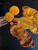 A hand holds two vibrant marigold flowers next to two exquisite Mulberry silk eye masks from Rimmba | Sustainable Clothing. One is the yellow Eco-Print Silk Eye-Mask with an elastic band, and the other features warm satin tones, all beautifully placed on a dark wooden surface.