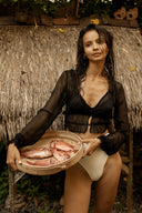 A woman with wet hair stands in front of a thatched hut, wearing a black Zena Silk Chiffon Top from Rimmba The Brand and white swimsuit bottoms, holding a wooden bowl filled with fish. Tropical foliage beautifully frames the scene.