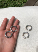 A hand elegantly holds Rimmba's Silver Baby Hoop earrings with a hammered beaten design against white fabric, while another pair rests beside on grass and wood.