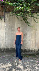 A person stands barefoot on a stone path, wearing a Kelly Strapless Silk Dress by Rimmba | Sustainable Clothing. Behind them, green vines and pink flowers enhance the textured concrete wall, dyed botanically for added elegance against the natural scene.
