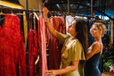 Two women are browsing dresses on a clothing rack in a boutique. One woman in a green dress closely examines a red garment, while the other in a blue dress looks at the clothes beside her. The store has warm lighting.