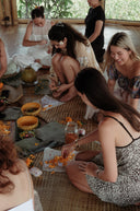 a group of people sitting together on the floor in a relaxed, natural setting. They are engaging in a hands-on activity involving flowers—mainly yellow petals and sappan wood —arranging or crafting them on pieces of cloth or leaves.