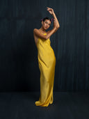 A model in the Mila Halter Silk Dress - Marigold by Rimmba | Sustainable Clothing poses gracefully against a dark textured backdrop, arms raised and head tilted, with flowers adorning her hair.