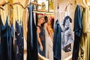 Two women stand close together in front of a round mirror in a clothing store, surrounded by hanging blue dresses. One woman wears a sheer navy dress, the other a white outfit, and they appear to be taking a mirror selfie.