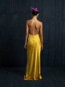 A woman with flowers in her hair stands against a dark, textured background, wearing the Rimmba Mila Halter Silk Dress in Marigold—a sustainable maxi dress featuring a halter-neck and open back.