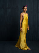 A woman stands barefoot against a dark backdrop, wearing the Rimmba Mila Halter Silk Dress in Marigold, with flowers in her hair and a serene expression.