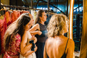 Four women stand closely together in a stylish clothing store, looking at dresses on a rack. One woman helps another adjust her dress while the others watch attentively. The setting is warmly lit and modern.