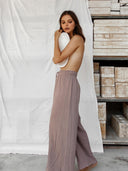 A woman with long brown hair stands sideways, topless, holding a white oval to her chest. She wears Rimmba The Brand’s Akasha 100% Cotton Pants in Lilac. Behind her is a white sheet and shelves with rectangular stone blocks.