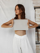 A woman wears Rimmba The Brand's Akasha Cotton Pants in white, holding a rectangular light gray tray against her bare upper body, standing before a white cloth backdrop and stacked boxes.