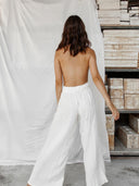 A woman with shoulder-length brown hair stands barefoot, facing away, wearing Rimmba The Brand's Akasha Cotton Pants - White with an elastic waistband. She is in front of a white sheet backdrop, wooden boxes stacked on metal shelves to the right.