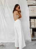 A woman with long brown hair stands barefoot on concrete, posing in front of a white backdrop with wooden boxes beside her. She wears Rimmba The Brand's Akasha Cotton Pants - White, featuring an elastic waistband and a loose fit.
