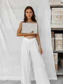 A woman with long brown hair and a neutral expression wears Rimmba The Brand’s Akasha Cotton Pants - White, featuring an elastic waistband. She stands before a white sheet, holding a closed laptop, with stacked boxes on shelves nearby.