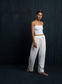 A woman stands barefoot against a dark backdrop, wearing the Akasha Cotton Pants in White by Rimmba The Brand and a strapless white smocked top. She has short hair with pink clips and a calm, relaxed expression.