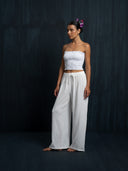 A woman barefoot against a dark textured background wears Rimmba The Brand’s Akasha Cotton Pants in white with a strapless top. Flowers adorn her hair as she gazes calmly to the side.