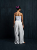 A woman stands barefoot facing a dark wall, wearing a white strapless top and Rimmba The Brand’s Akasha Cotton Pants in white. Her hair is tied up with purple flowers.