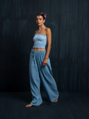Barefoot woman wearing a baby indigo cotton strapless top and long cotton pants against a dark background.