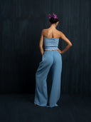 Barefoort woman wearing a baby indigo cotton strapless op and long cotton pants is facing a dark background.
