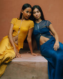 two women sitting closely together on a ledge against a warm, earthy-toned wall. They are leaning slightly toward each other, creating a relaxed and intimate pose.

One woman is wearing a mustard-yellow outfit with a semi-sheer top and a long skirt, while the other is dressed in a deep blue outfit with a similar lightweight, semi-transparent top and matching skirt.