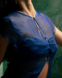 a close-up of a person’s upper torso, focusing on an Alila Silkchiffon blouse indigo. The fabric is lightweight and semi-transparent, revealing subtle details of the body underneath. The top features short sleeves and a vertical row of small buttons down the front, with a narrow opening at the chest that adds a delicate, stylish touch.