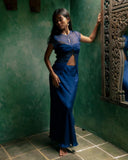 a woman standing gracefully in a corner of a room with textured green walls and tiled flooring. She is wearing an indigo, floor-length outfit consisting of a fitted skirt and a sheer, short-sleeved top.She stands barefoot, with one hand gently touching an ornately carved wooden window frame, while the other rests naturally by her side.