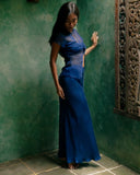 A woman standing indoors against a textured green wall. She is wearing Silkchiffon Blouse indigo and elegant Aman bias silk skirt. Her long dark hair falls over one side of her shoulder, and she is looking down with a calm and sligthly shy expression.