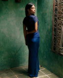 A woman standing indoors, facing sligthly away from the camera. She wearing Alila Silkchiffon blouse indigo and matching Aman bias skirt indigo.