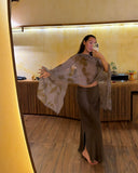 Karunia Fischer's taking a mirror selfie in a warmly lit room with wooden flooring and modern interior decor. She is posing gracefully with one arm extended outward, showcasing a flowing silkchiffon scarf ecoprint grey. She pairs the elegant top with a silk satin aman mud skirt, creating a sophisticated and earthy look.