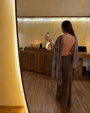 A woman is taking a mirror selfie in a warmly lit, minimalist room with wooden flooring and soft ambient lighting. She is wearing an elegant backless aman bias silk skirt mud paired with a sheer ecoprint silk chiffon scarf draped loosely over her arms. Her long dark hair falls naturally down her back, adding to the sophisticated and graceful atmosphere of the photo.